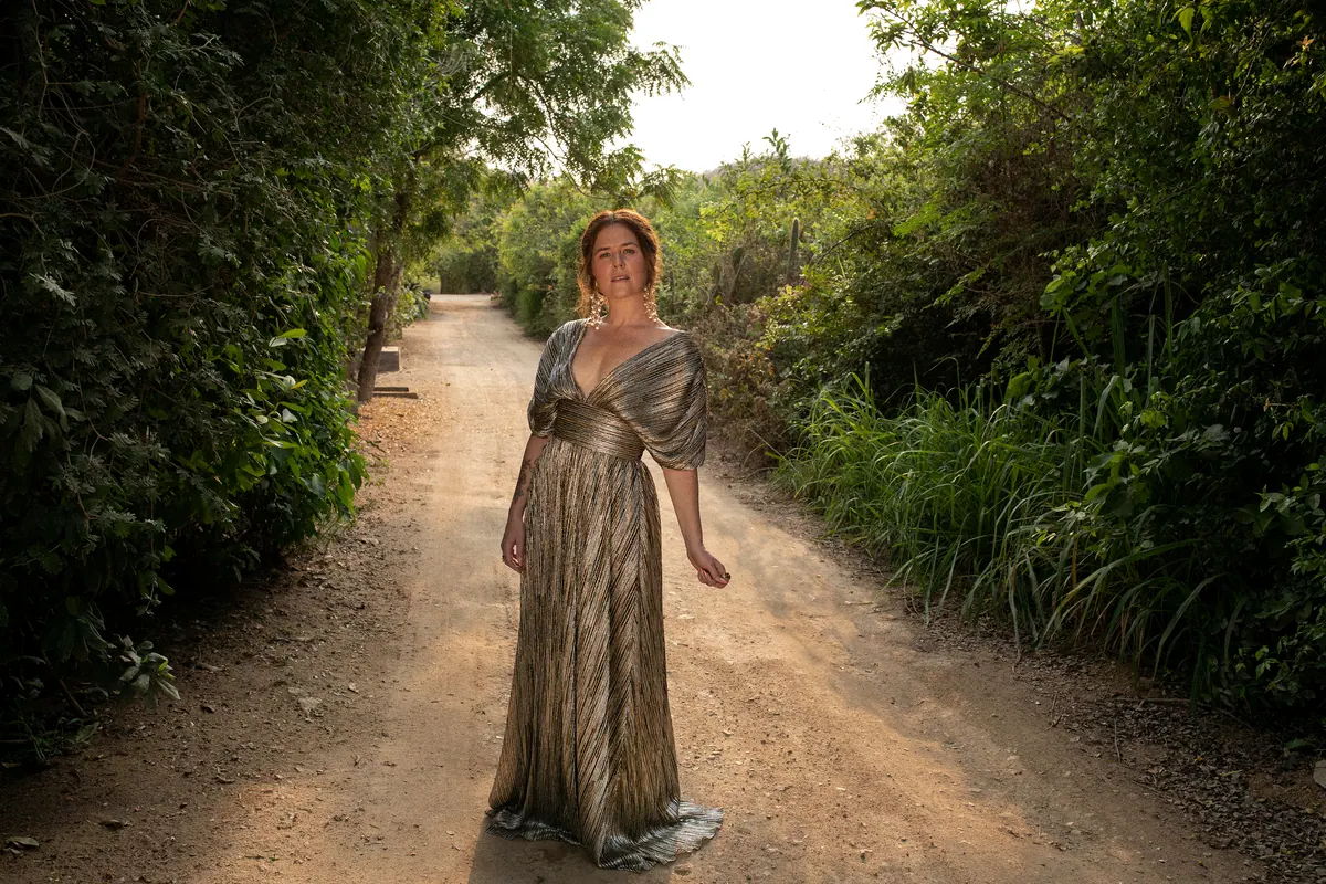iskwē standing on a dirt path between green trees in a metallic gold gown