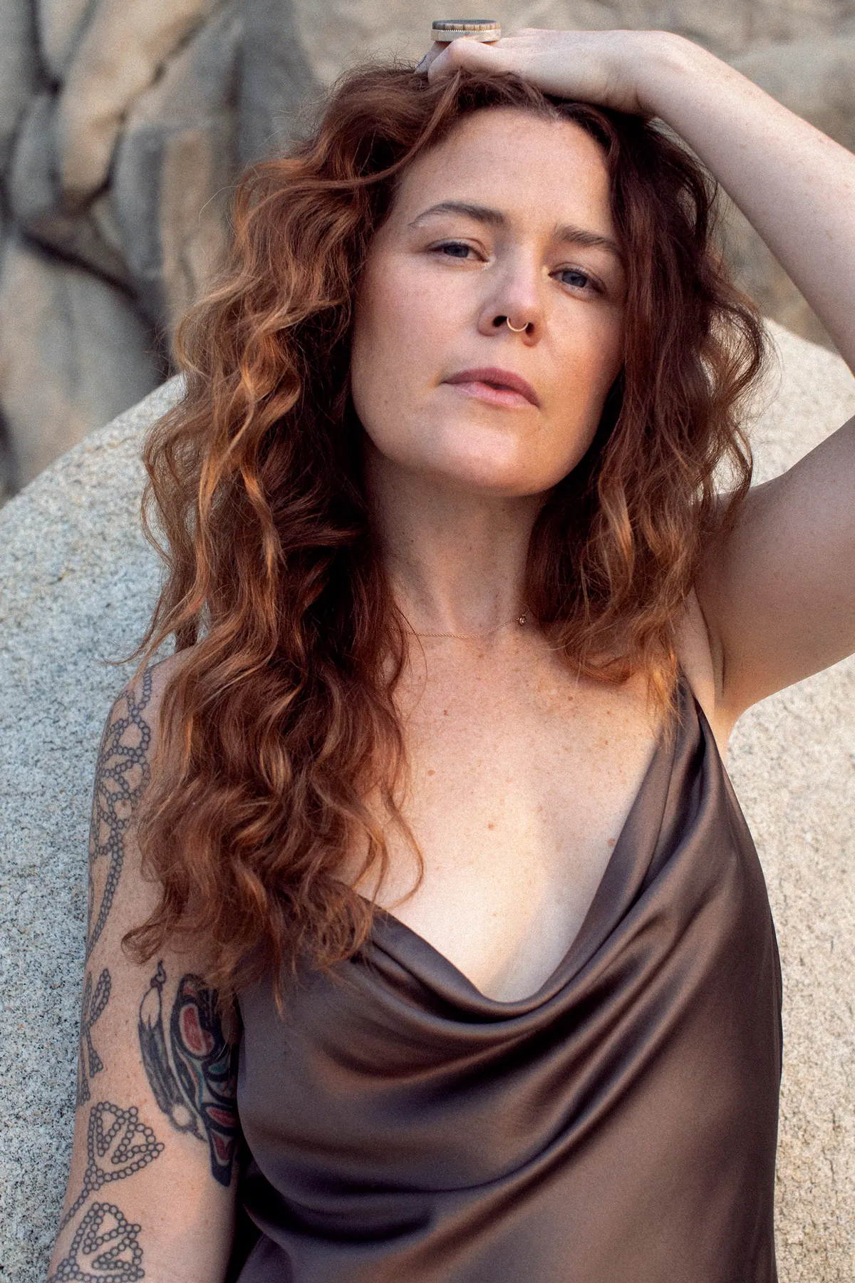 iskwē in a bronze satin dress, leaning against a rock with one hand on her head